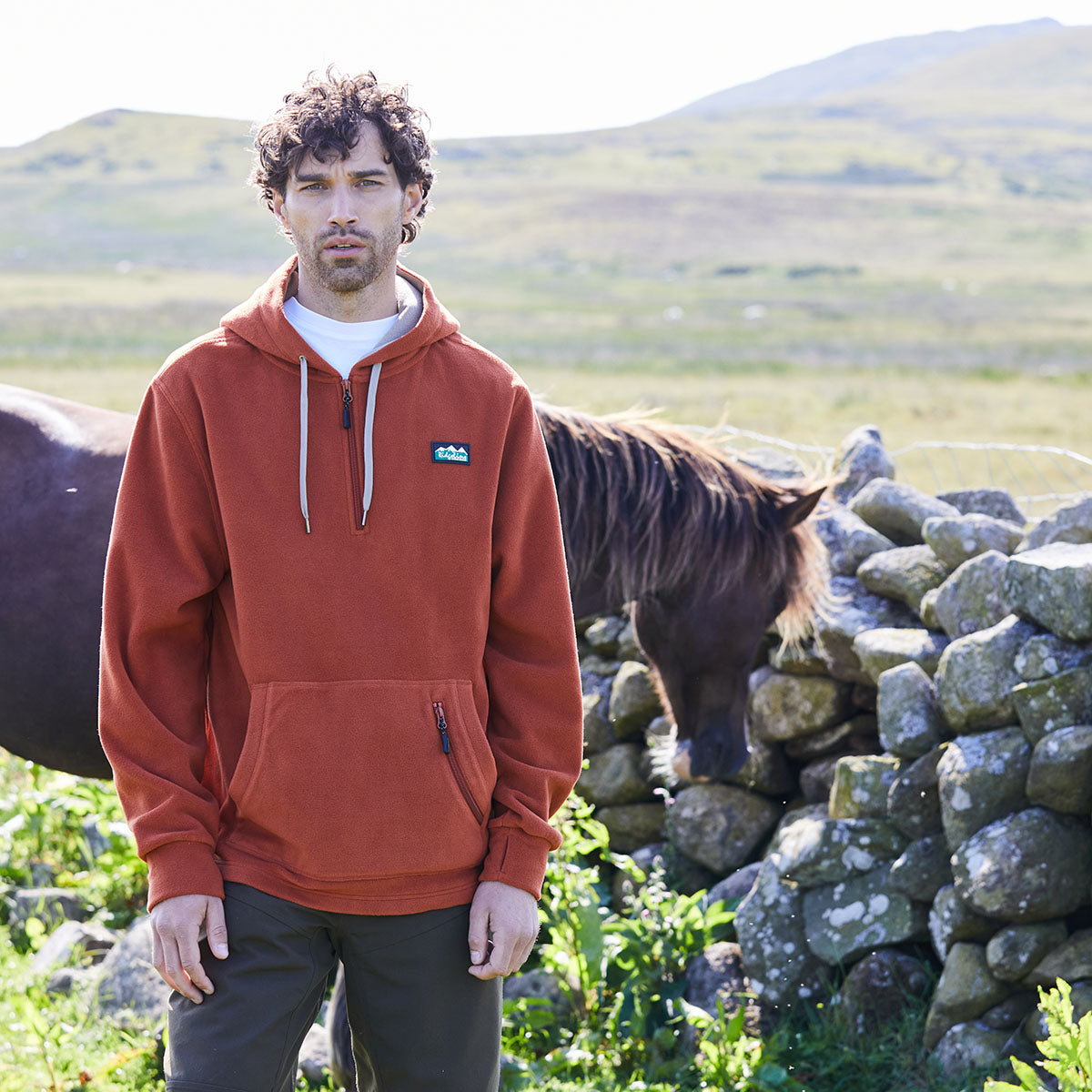 Man wearing an orange hoodie with Ridgeline Clothing logo on chest standing next to a horse in a field with stone walls and hills in the background.