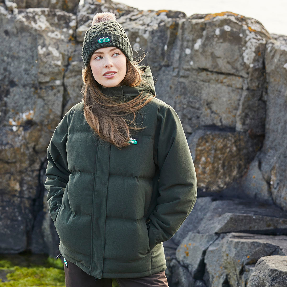 A person standing outdoors in front of a rugged stone wall, wearing a dark green padded jacket and a knitted beanie hat with a pom-pom. The jacket has front pockets with the person's hands tucked inside. Long hair is visible beneath the hat. The background features large gray rocks and a small patch of green grass, suggesting a cool-weather rural or coastal setting. At the bottom of the screenshot, a website editing overlay displays the text “Click image to set focal point.”
