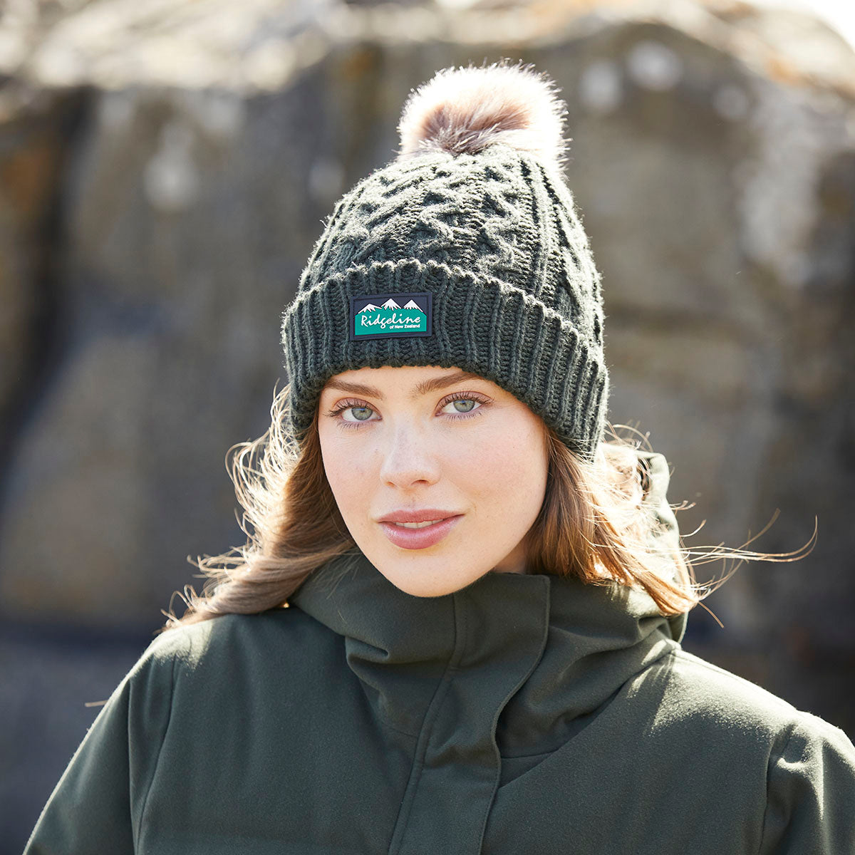 Lifestyle image of a person wearing an olive green cable-knit beanie with a deep ribbed cuff and a large faux-fur pom-pom on the crown. A Ridgeline of New Zealand logo patch is attached to the front cuff. The beanie features a textured cable-knit pattern throughout and is paired with an olive green outdoor jacket. The photograph is taken outdoors with a softly blurred rocky background and natural daylight, highlighting the beanie's warm knit construction, comfortable fit, and stylish cold-we