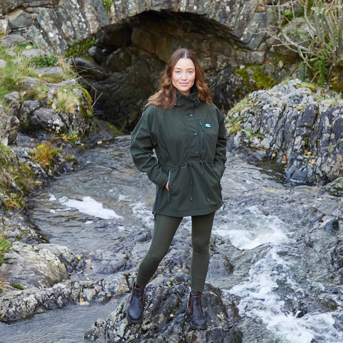 woman wearing a A green Monsoon II Classic Smock with a hood, featuring a front zip and multiple pockets. Standing on a rock in the middle of a stream 
