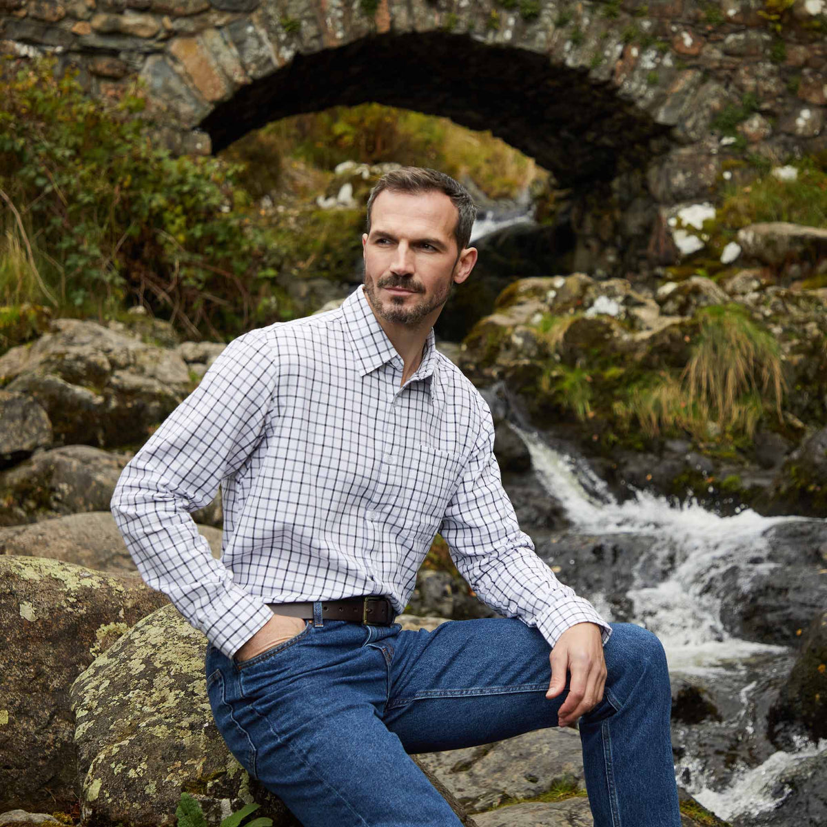 Man sitting on rocks by a stream with a stone bridge in the background, wearing a checked shirt