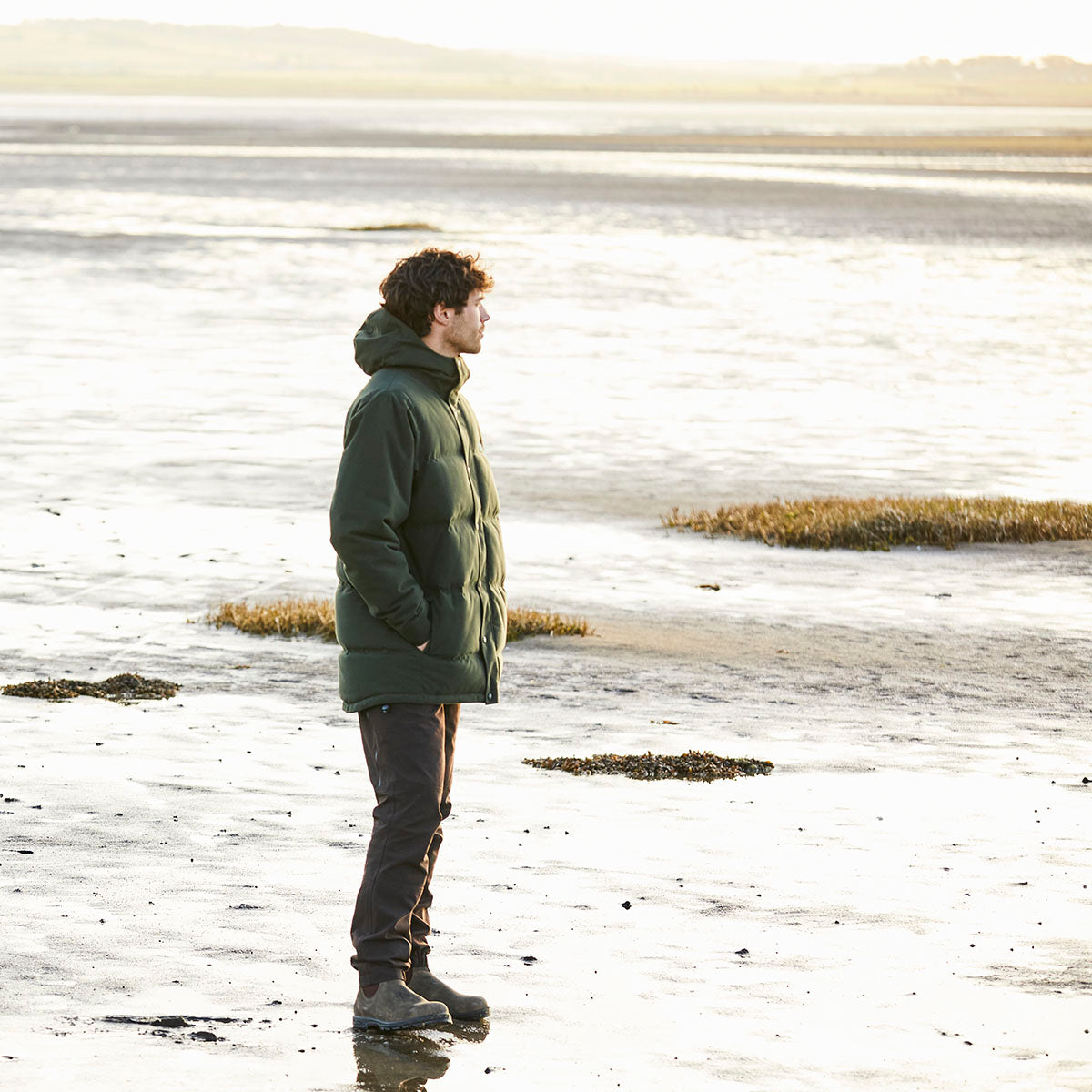 Dark green padded hooded jacket, brown trousers and dark ankle boots, side view worn outdoors on sandy shoreline with water and grassy patches in background