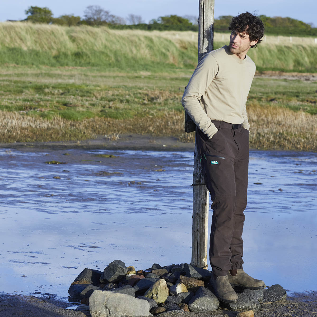 Ridgeline brown woven fabric belt worn with trousers by model, outdoor setting by a body of water.