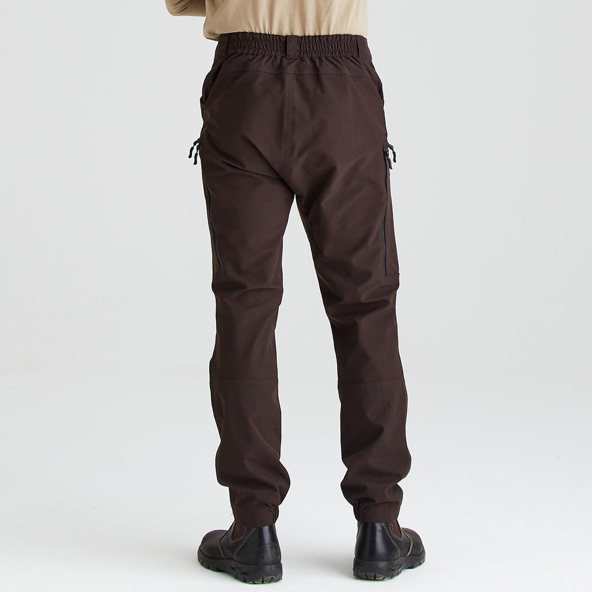 Rear view of dark brown work trousers worn with black work boots, displayed against a light grey studio background. The trousers feature an elasticated waistband, straight-leg fit, articulated knee panels, and vertical zippered pockets positioned on the outer back of each leg. A beige long-sleeve top is partially visible above the waistband