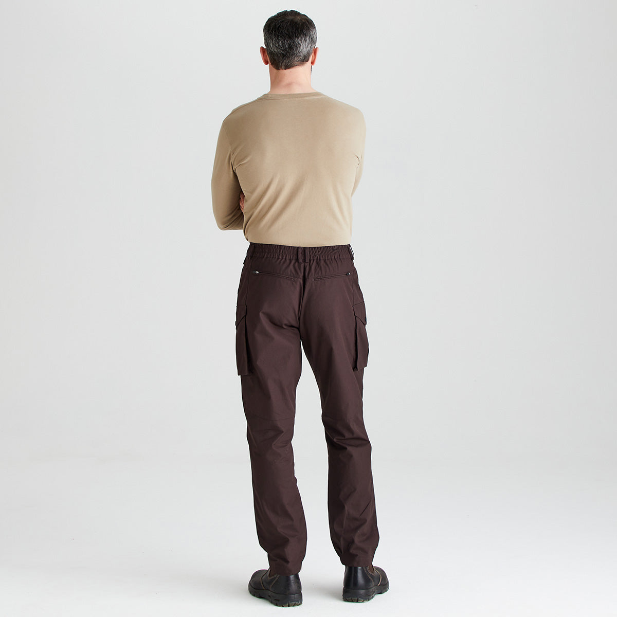 A man viewed from behind wearing dark brown cargo pants, a beige long-sleeve shirt, and black shoes, standing against a light gray studio background.