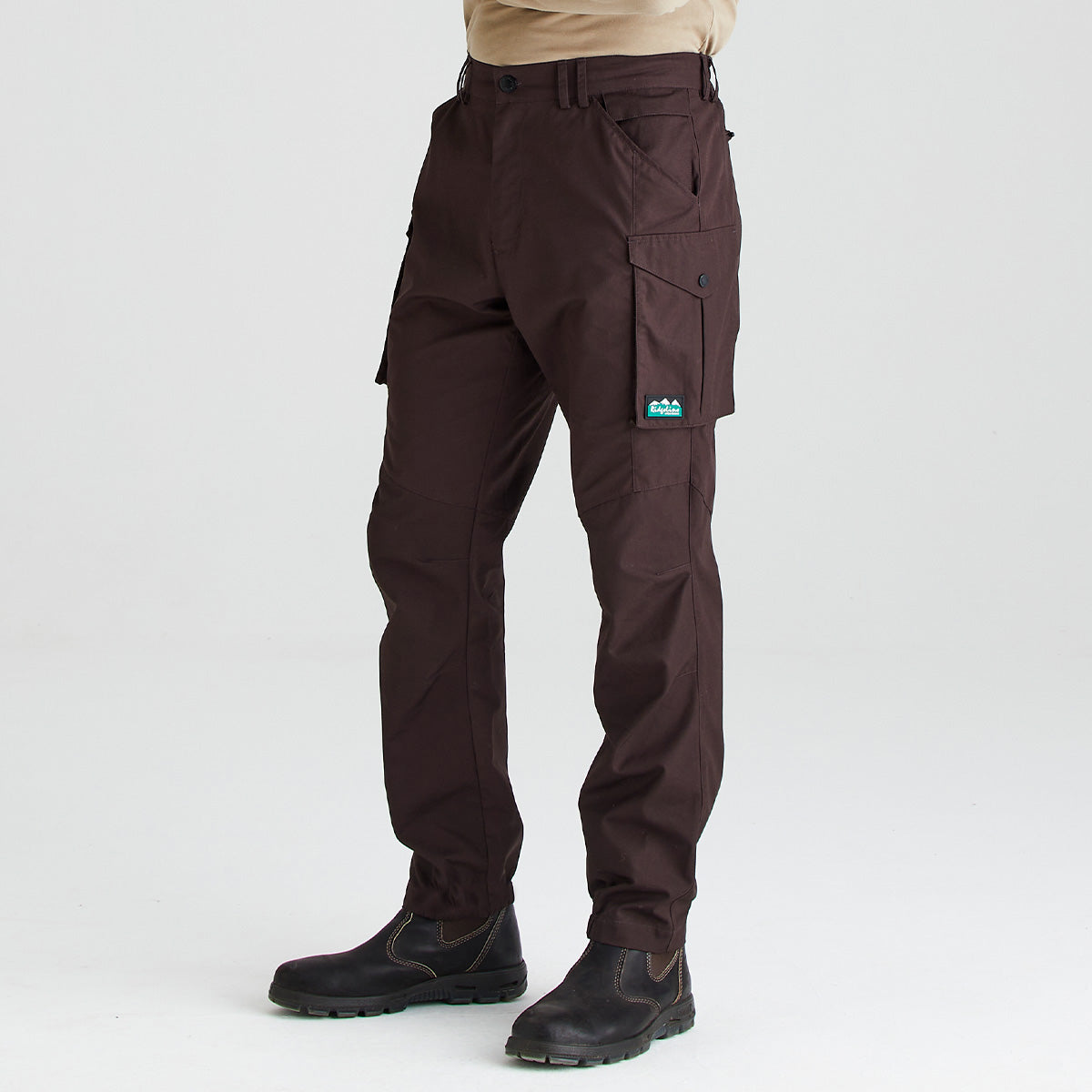 Dark brown cargo pants worn by a model, paired with black work boots and a beige shirt, shown from the waist down on a light gray studio background.