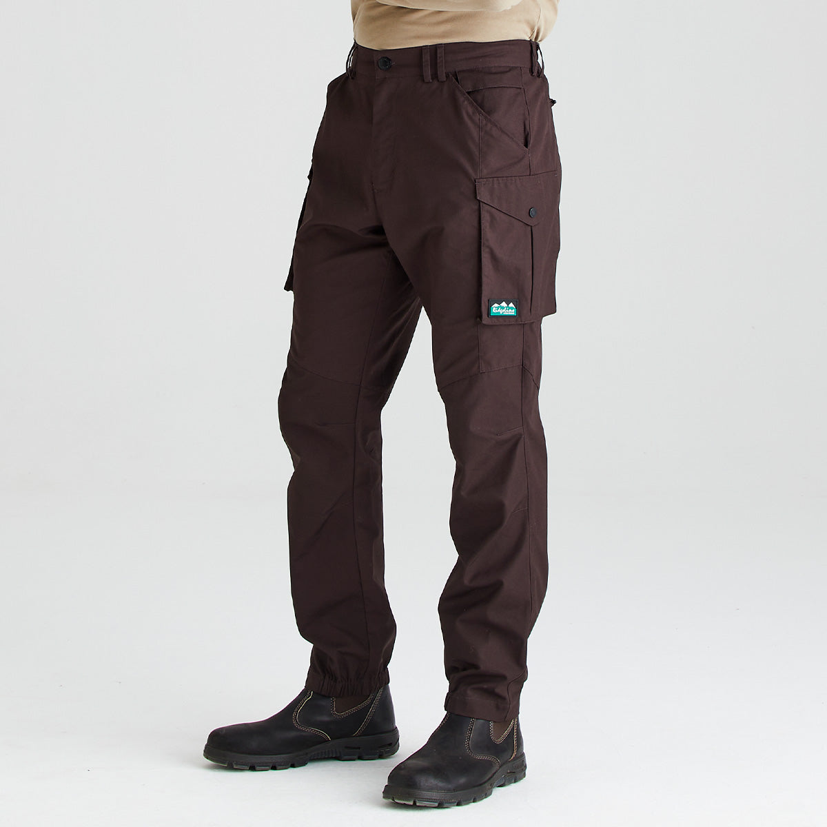 Three-quarter side view of dark brown cargo work trousers featuring multiple utility pockets, including a large button-fastened cargo pocket on the thigh, belt loops, and elasticated ankle cuffs. The trousers are shown with black work boots against a light grey studio background.