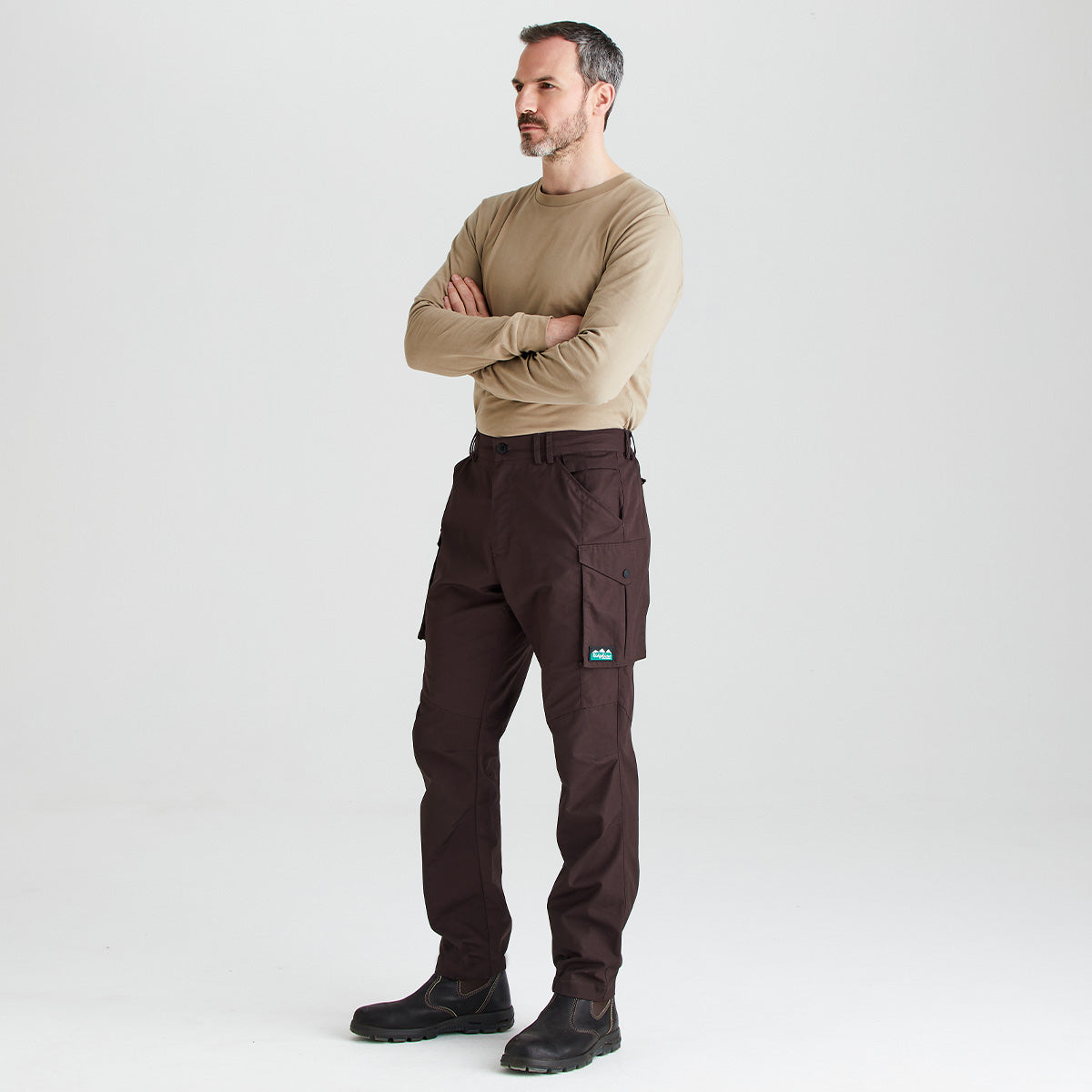 A man modeling dark brown cargo pants with a tan long-sleeve shirt and black boots, standing against a light gray studio background.