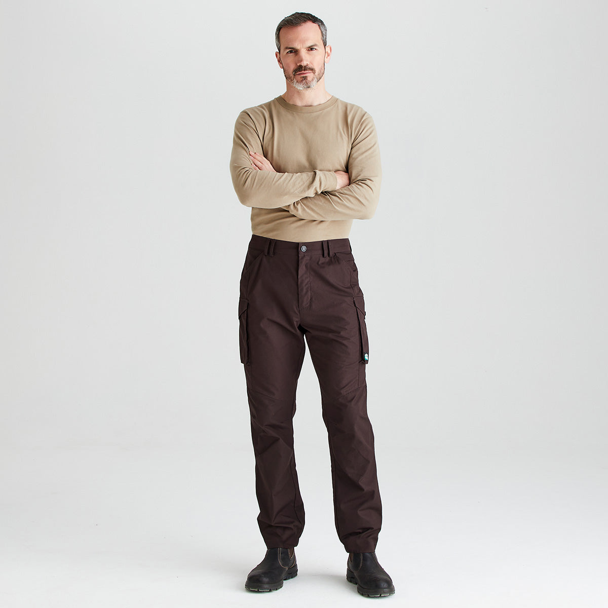 A man stands with arms crossed wearing dark brown cargo pants, a beige long-sleeve shirt, and black boots against a light gray studio background.