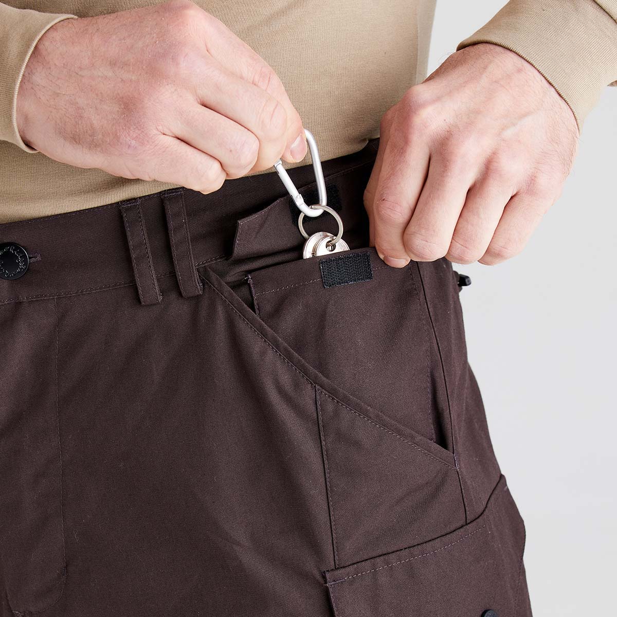 Close-up of dark brown cargo trousers showing a utility pocket at the waistband, with a silver carabiner clipped to a fabric loop and a retractable ID holder stored inside the pocket. The trousers feature belt loops and multiple storage pockets.