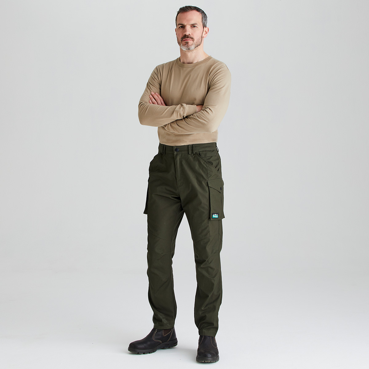 A man stands with arms crossed wearing olive green cargo pants, a beige long-sleeve shirt, and black shoes against a light gray studio background.