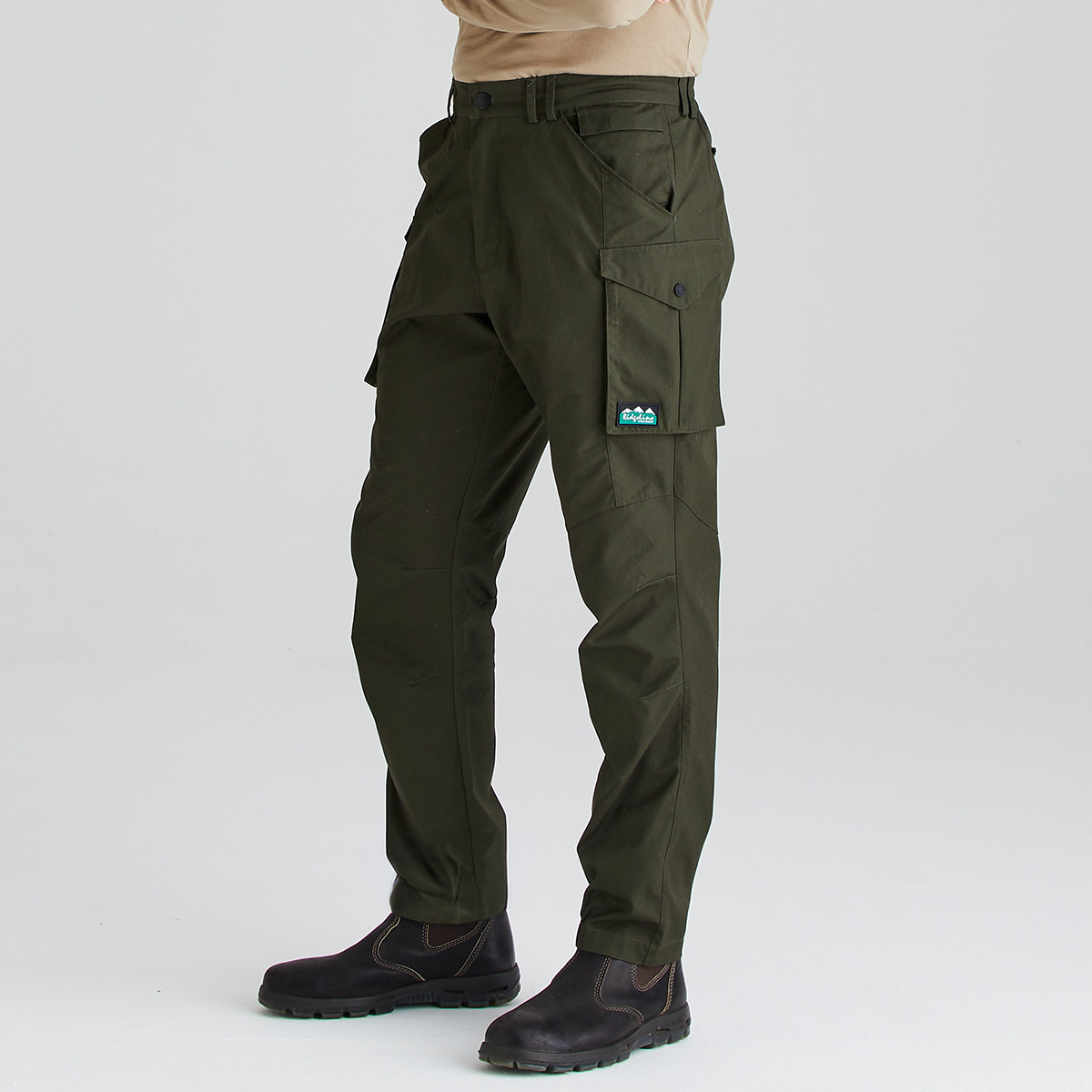 A person wearing dark olive green cargo pants with a small blue and white logo patch on the pocket, paired with dark brown boots against a light gray studio background.