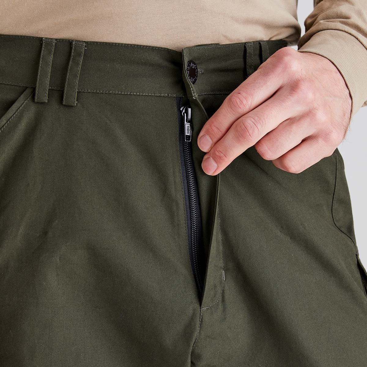 Close-up view of olive-green cargo work trousers showing the front waistband and fly. A hand is pulling back the fly cover to reveal a black zip fastening beneath a buttoned waistband with belt loops. The image highlights the trousers' secure zip-fly closure and durable workwear construction against a neutral studio background.