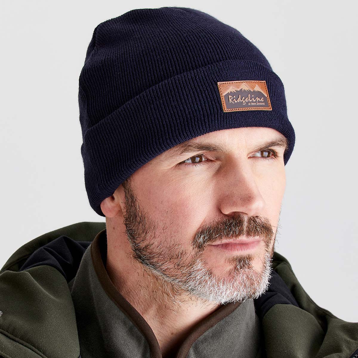 Close-up product image of a person wearing a navy blue ribbed knit beanie with a folded cuff. A brown faux-leather patch on the front displays the text “Ridgeline” with a mountain graphic and the words “of New Zealand.” The beanie is shown against a light grey background, highlighting the texture and fit of the hat.

