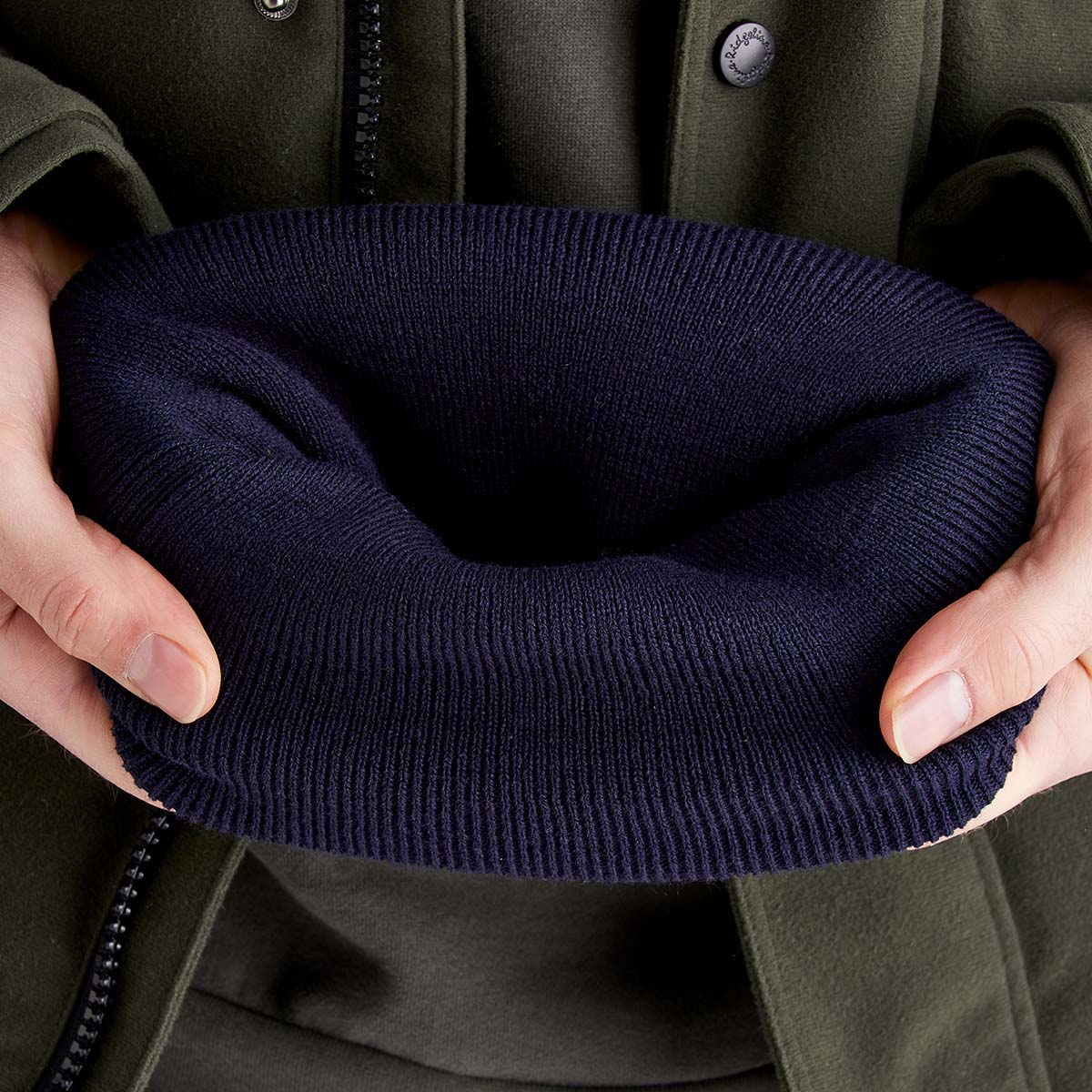 Close-up view of a navy blue knitted beanie being held open with both hands to show the interior and thickness of the fabric. The ribbed knit texture is clearly visible, and the beanie appears soft and stretchy. The person holding the hat is wearing a dark green zip-front jacket, with the jacket partially visible in the background. The image emphasizes the beanie's construction, depth, and warm knit material.
