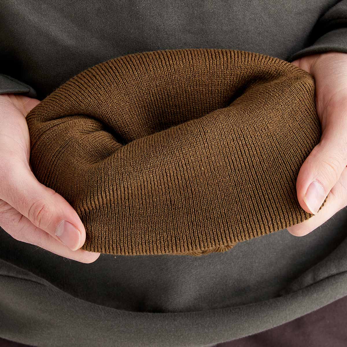 Close-up image of a greenish-brown ribbed knit beanie being held open with both hands to display the interior shape and thickness of the material. The fine-knit fabric and stretchy construction are clearly visible, emphasizing warmth and comfort.  The image highlights the beanie's soft texture, durability, and insulated design.
