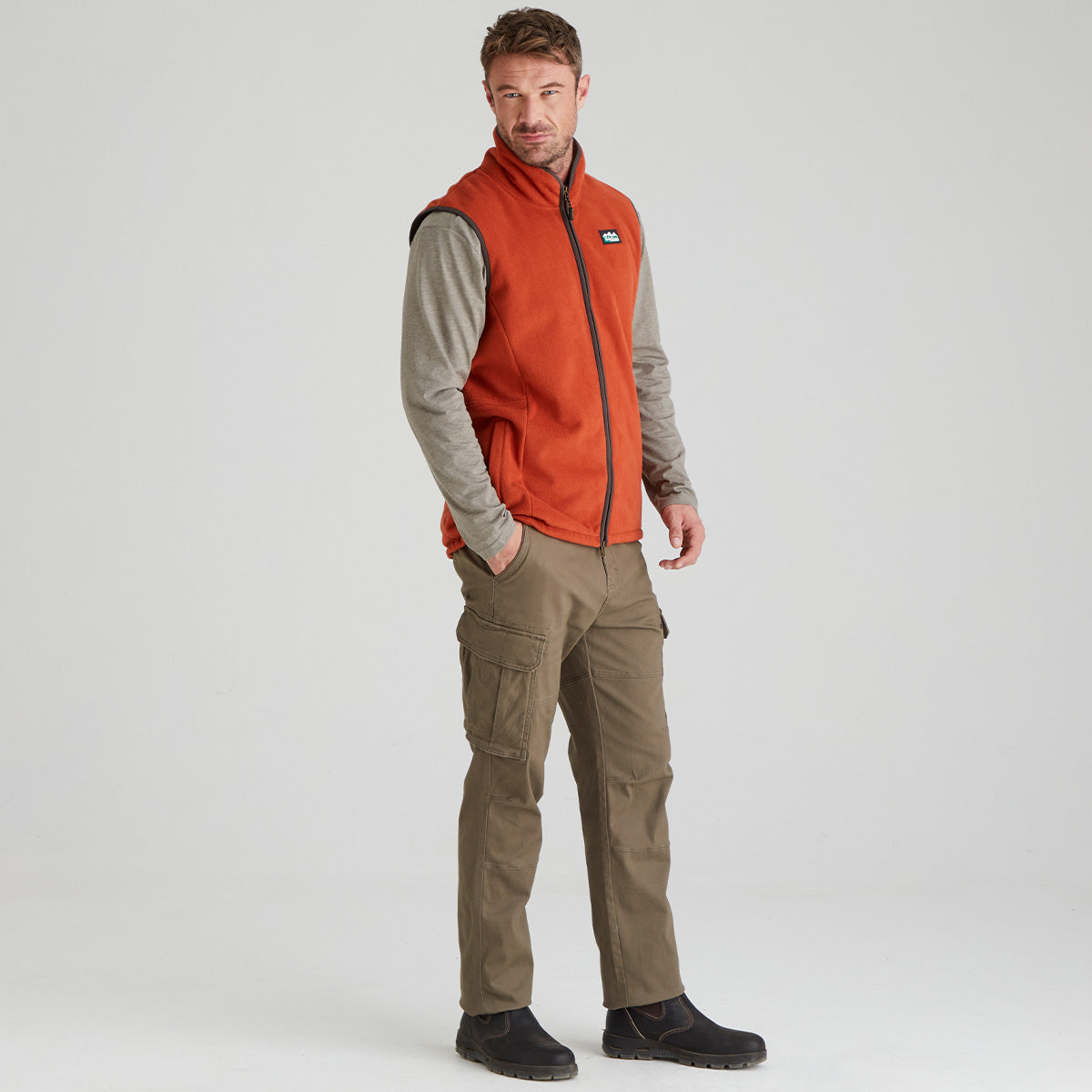 Man wearing an orange fleece vest over a grey shirt with brown pants on a light grey background
