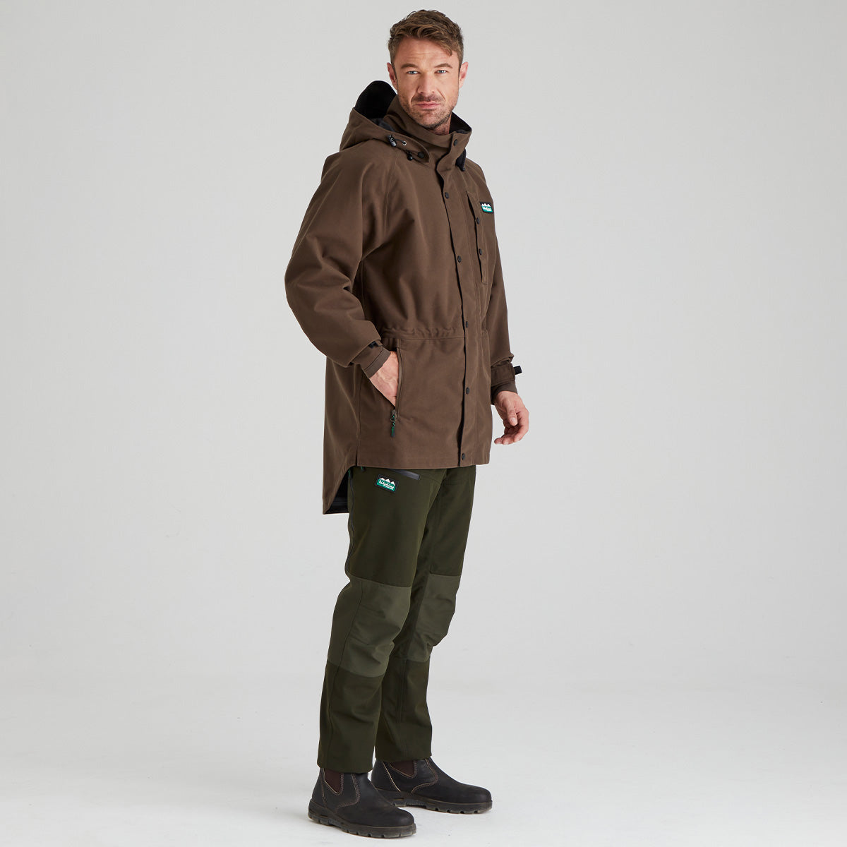 Man wearing a brown waterproof jacket and green pants on a grey background