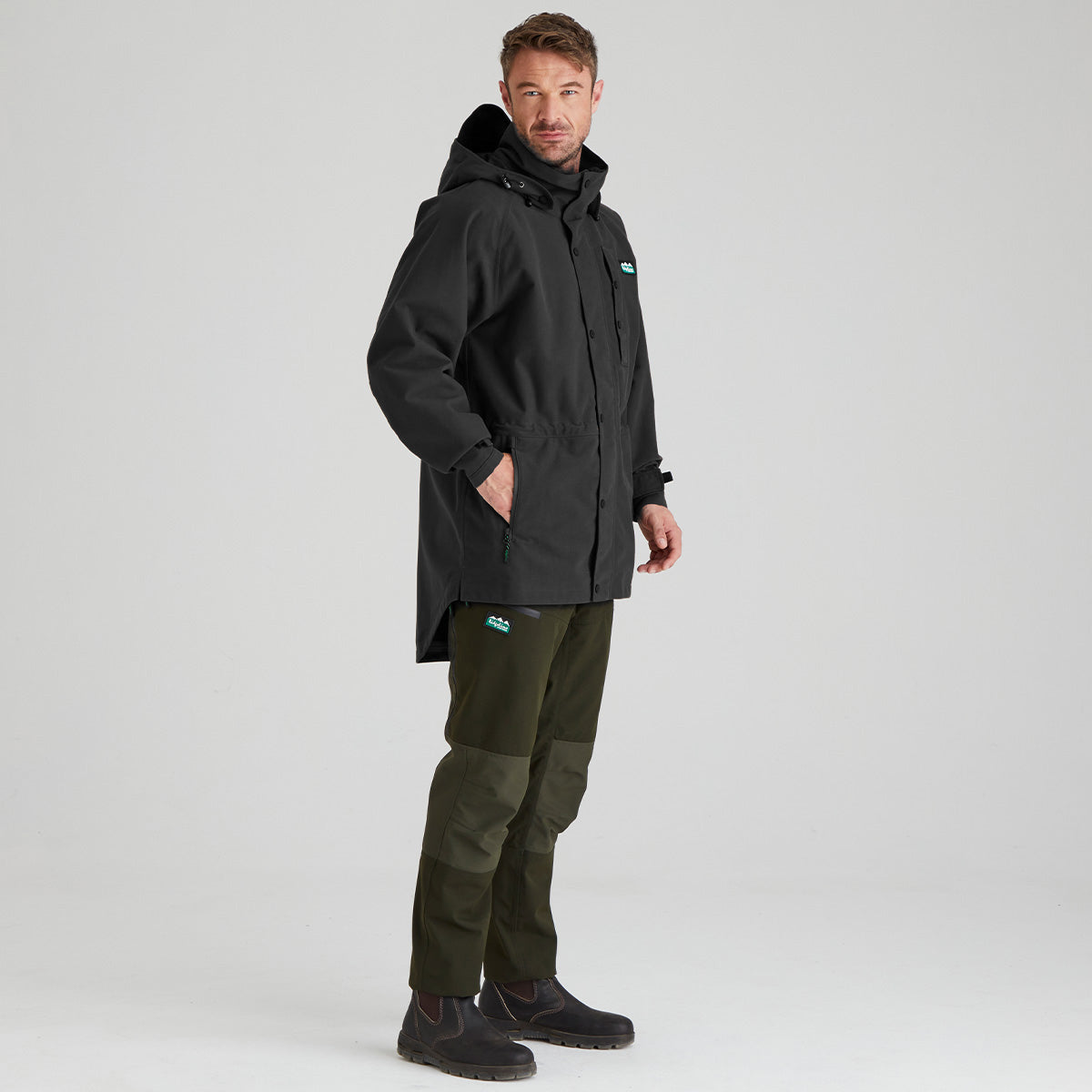 A man modeling a black hooded technical jacket and olive green pants against a light gray studio background.
