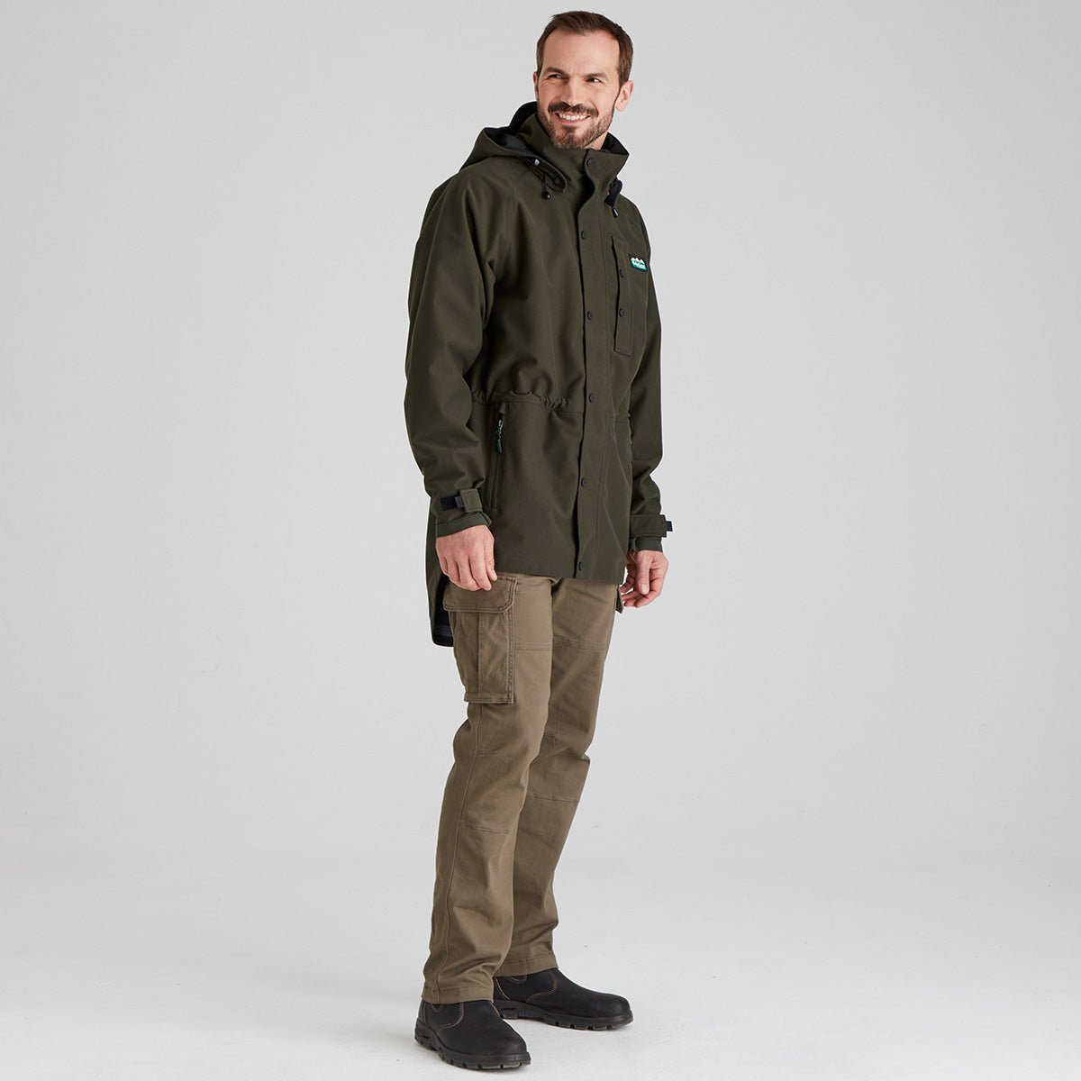 Man wearing a green waterproof jacket and brown pants on a grey background