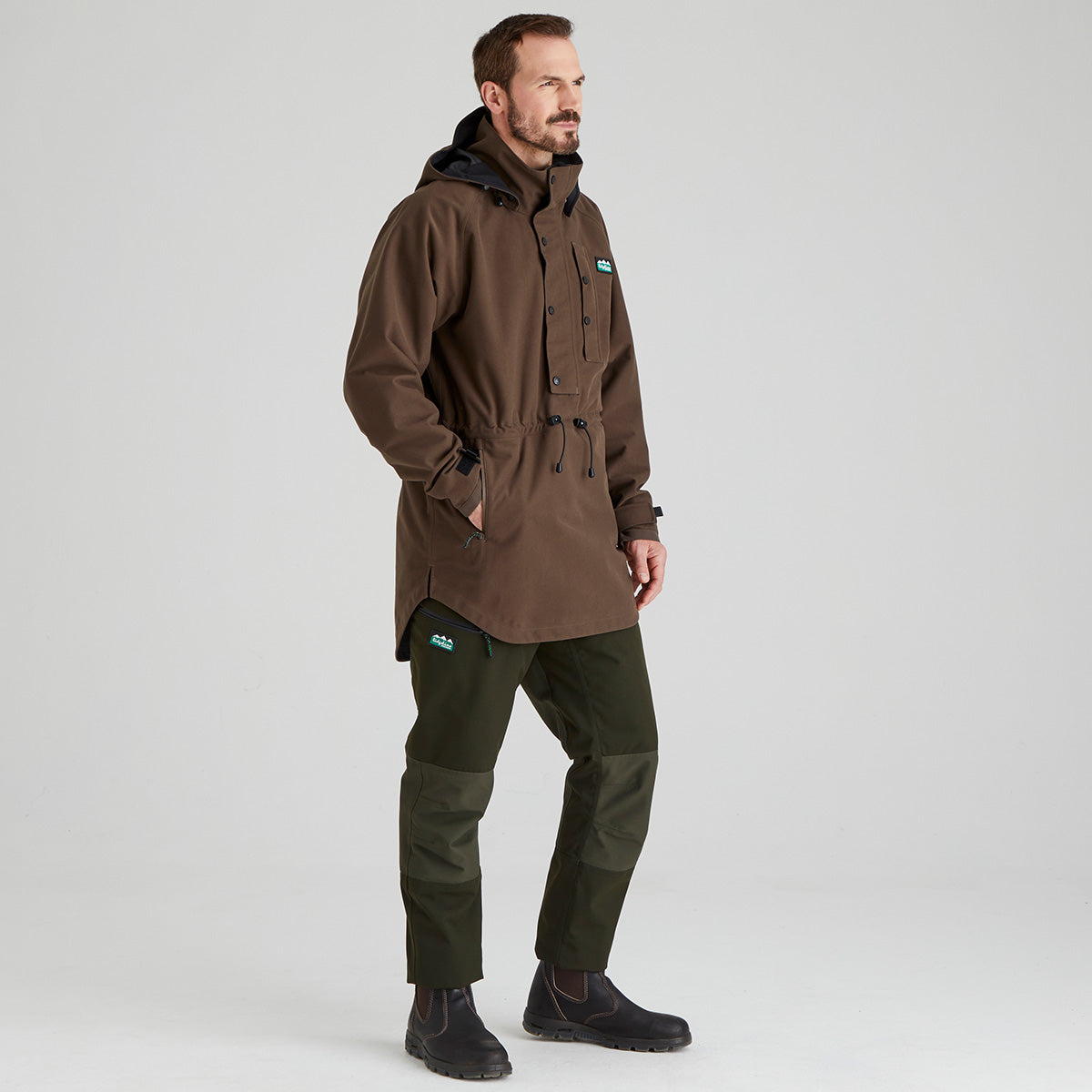 Man wearing a brown waterproof jacket and green pants on a light grey background