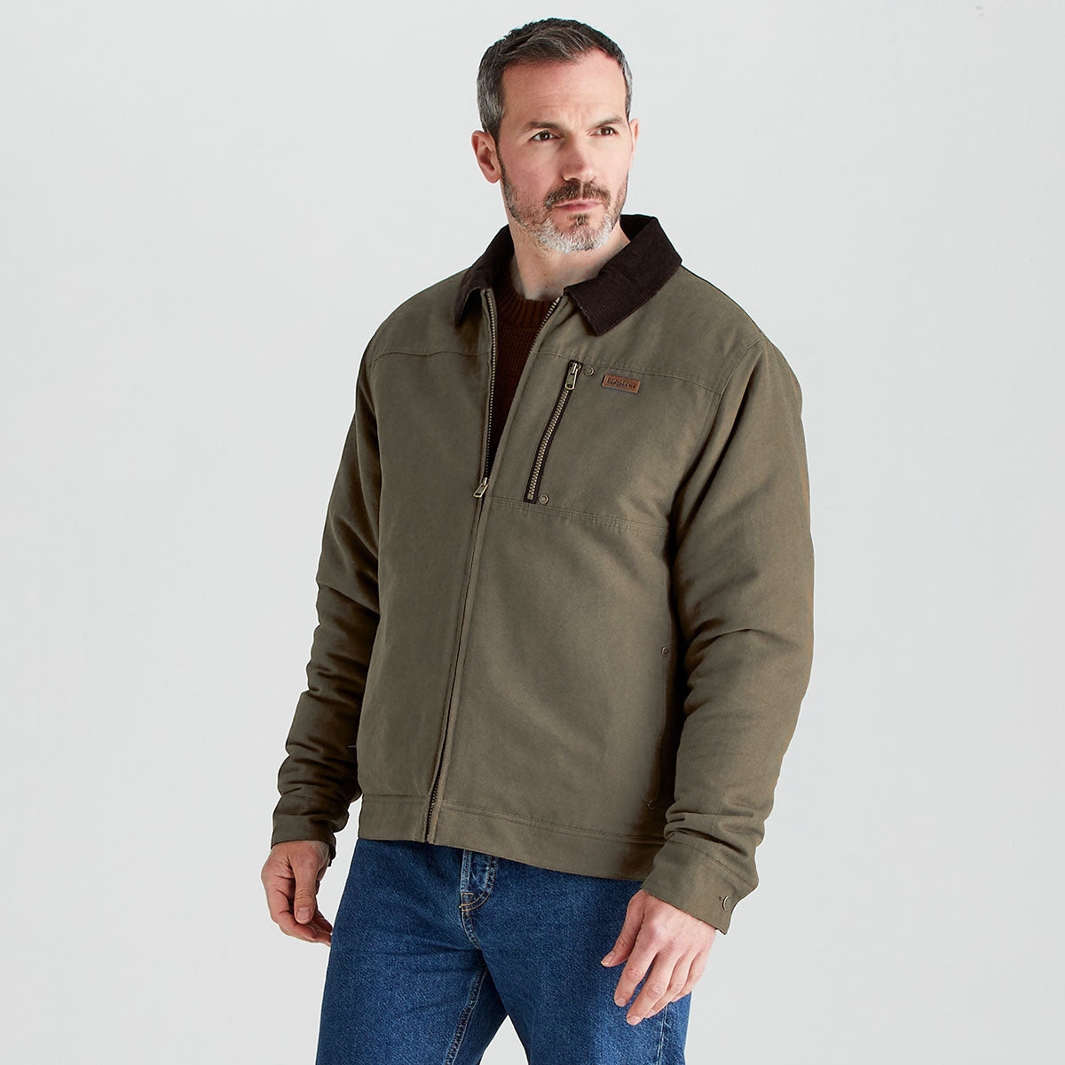 Person modeling an olive-green zip-front jacket with a dark brown contrast collar and vertical zip chest pocket, layered over a brown knit sweater and blue jeans, standing against a light gray studio background.
