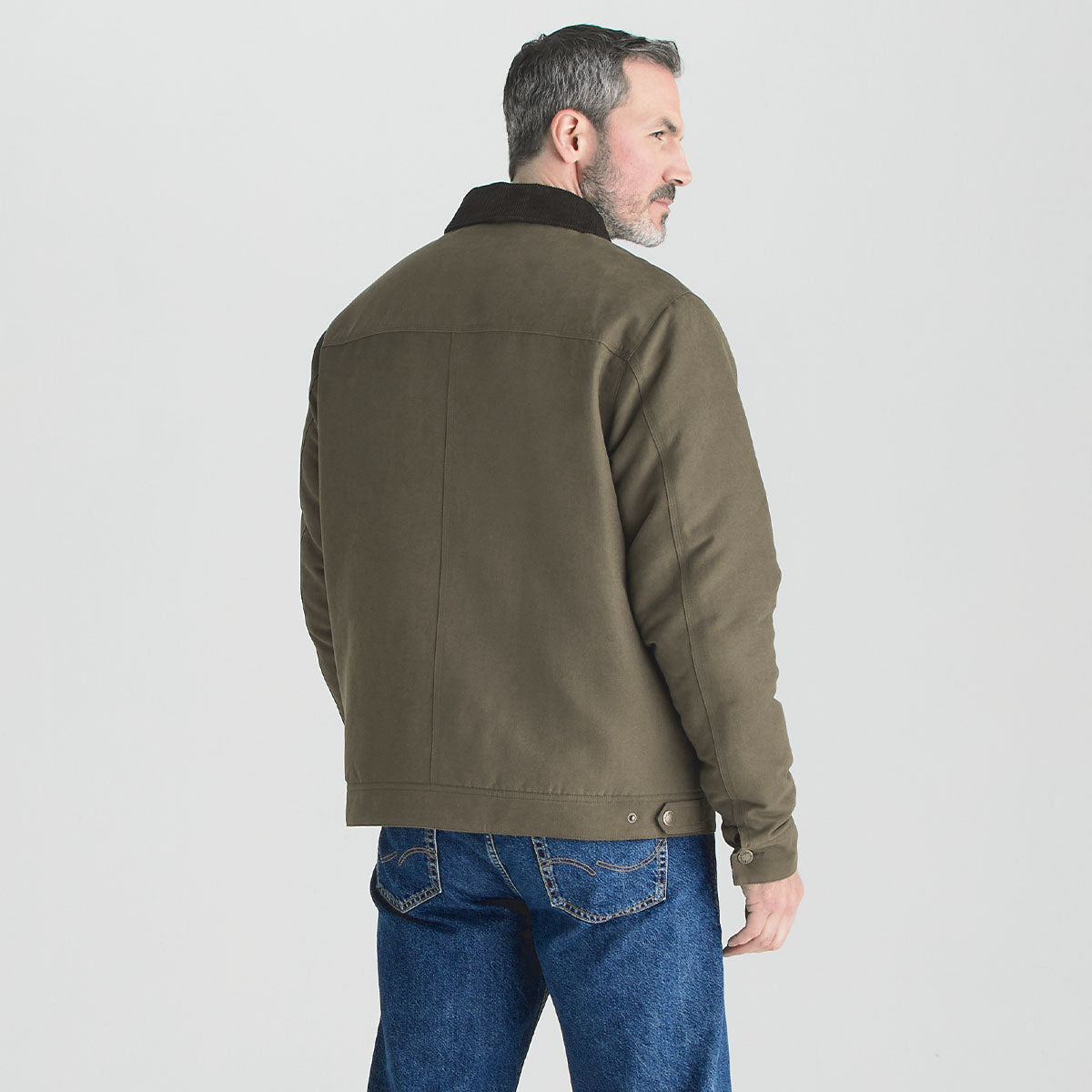 Rear three-quarter view of a person wearing an olive-green jacket with a dark brown contrast collar. The jacket is styled with blue jeans and shown against a plain light gray studio background, highlighting the back panel design, hem adjusters, and sleeve cuff details.
