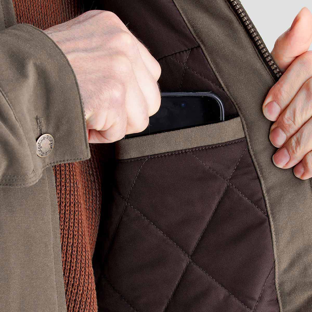 Close-up of the inside of an olive-green jacket showing a quilted brown lining and an internal slip pocket sized for a smartphone. A hand is placing a phone into the pocket, highlighting the jacket's storage feature, while the sleeve cuff and front zipper are visible in the frame.

