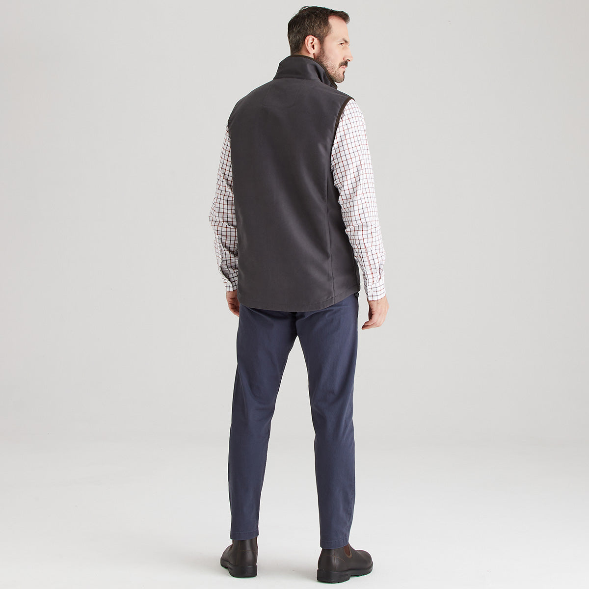 Man wearing a grey vest over a checkered shirt with navy pants on a light grey background
