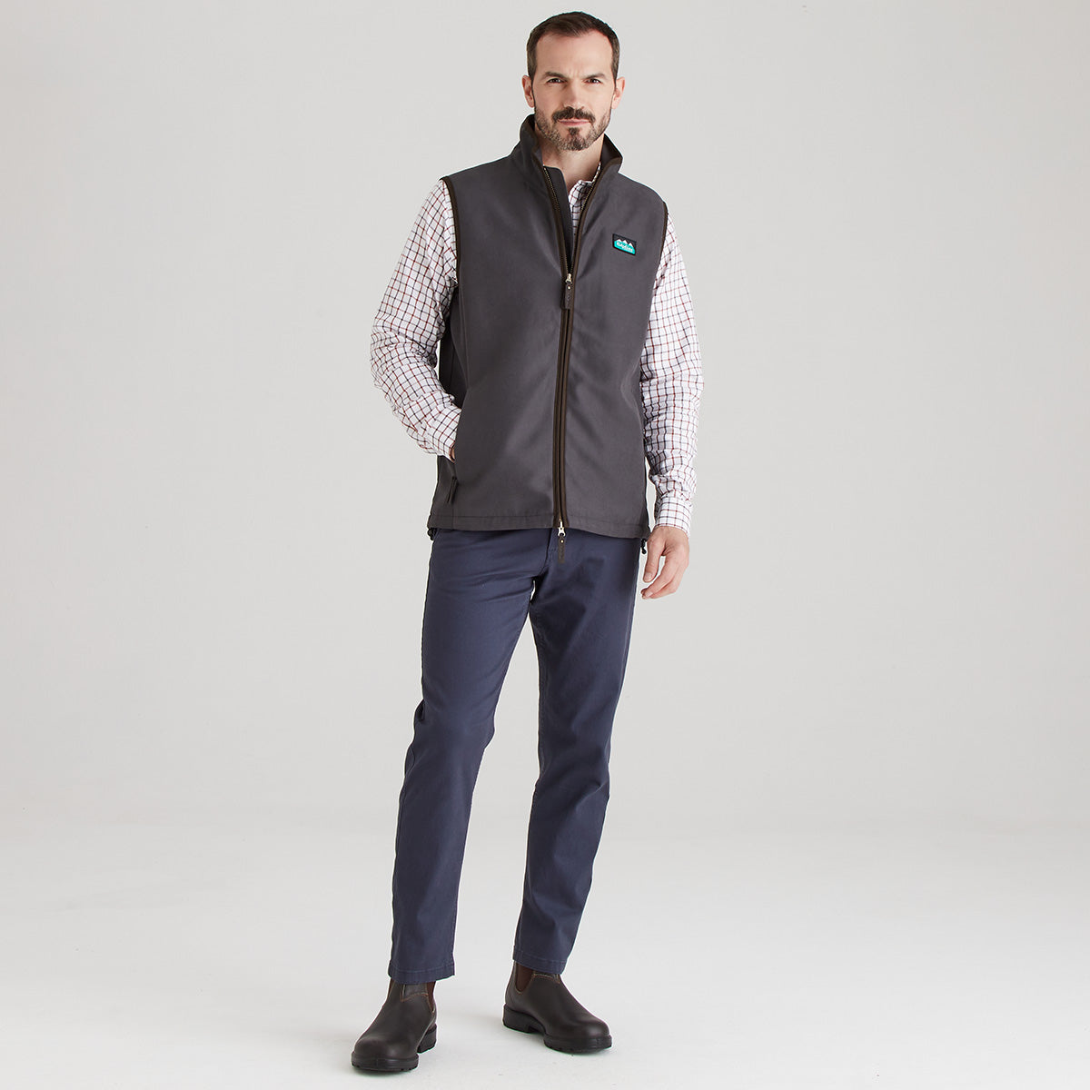 Man wearing a grey vest over a checkered shirt with a visible brand logo on a light grey background