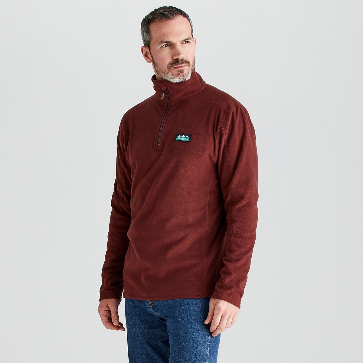 Person standing against a light gray studio background wearing a burgundy quarter-zip ridgeline fleece pullover and blue jeans. The fleece has a high collar with a short zipper and a small rectangular logo patch on the left chest. The person is shown from mid-thigh up, facing slightly to the side with one hand visible at the hip.
