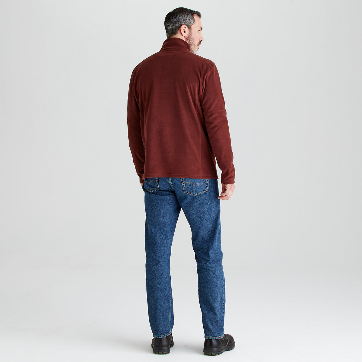 Rear view of a person standing against a light gray studio background wearing a burgundy quarter-zip fleece pullover, blue jeans, and dark shoes. The fleece features a high collar and long sleeves, with a plain back design. The person is shown full length, turned slightly to one side, displaying the back fit and length of the garment.
