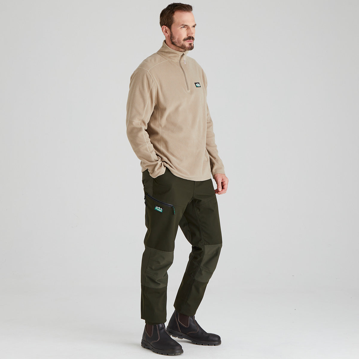 Man wearing a beige fleece pullover  and green pants on a light grey background