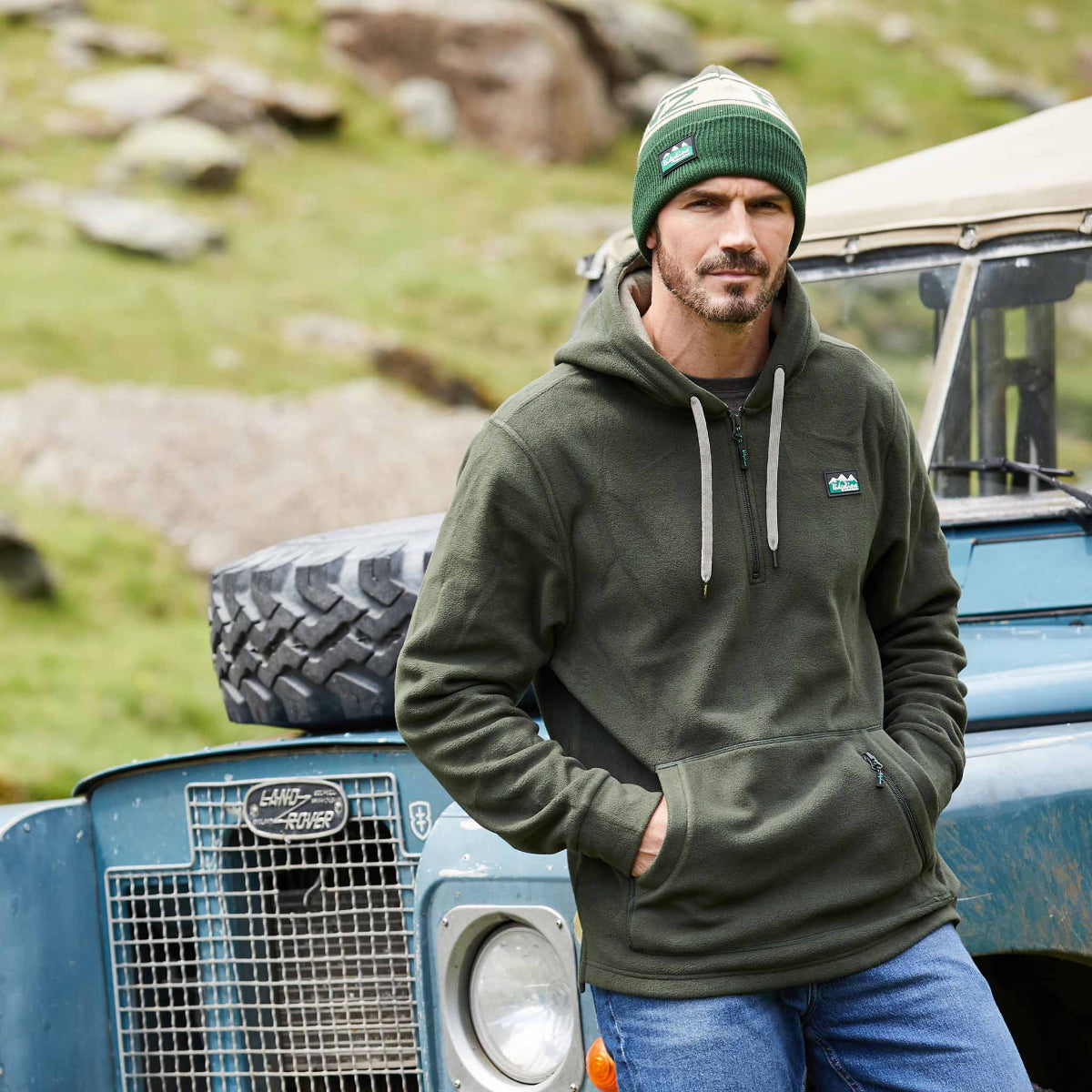 Man wearing a green fleece hoodie with a hood, 1/4 zip front, and thumb-loops at the cuffs. It features a front kangaroo pocket and a secondary zippered pocket. Ridgeline Clothing Logo on chest. Leaning on a car with Rocks in the background