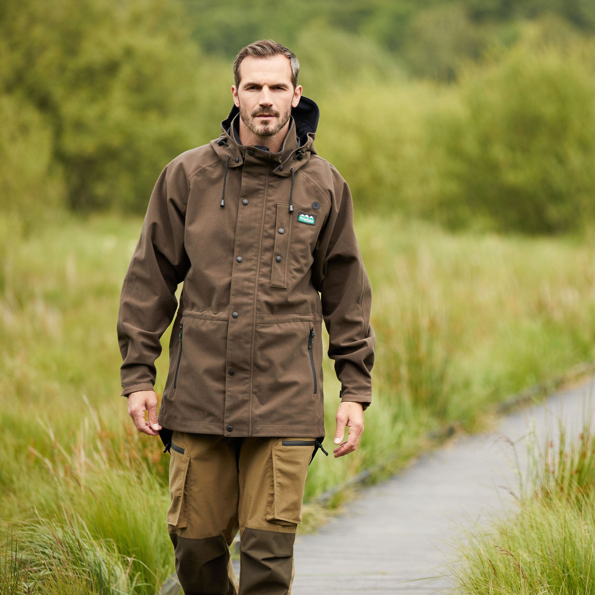 man wearing A brown-coloured Ridgeline monsoon waterproof Jacket with a detachable hood, zip front, and multiple pockets. walking through a walkway through grass