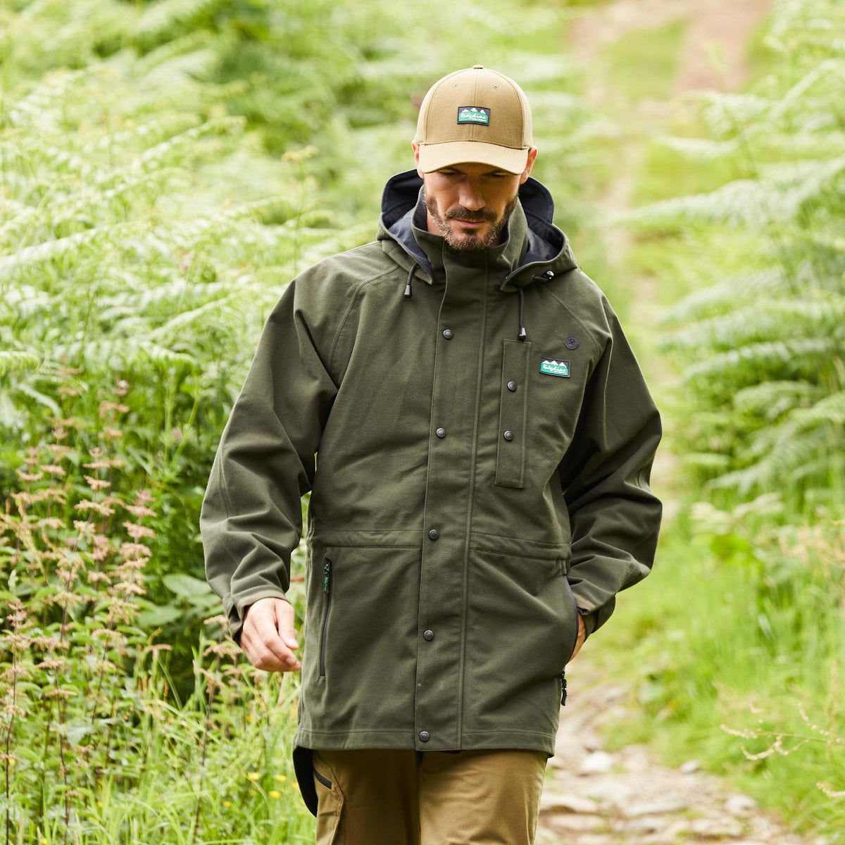 Man walking through walkway with high plants wearing a dark green-coloured Ridgeline Monsoon waterproof Jacket with a detachable hood, zip front, and multiple pockets. 