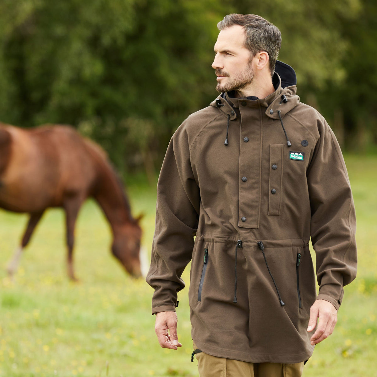 Man wearing a brown jacket standing in a field with a horse in the background