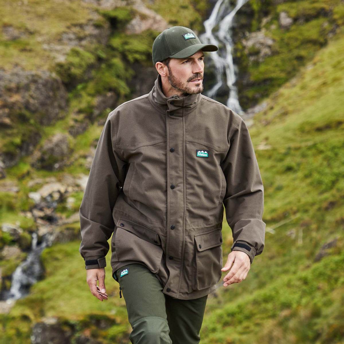 man wearing a brown coloured Ridgeline Torrent III Jacket with a detachable hood, zip front, and multiple pockets in an outdoor setting with a stream running in the background