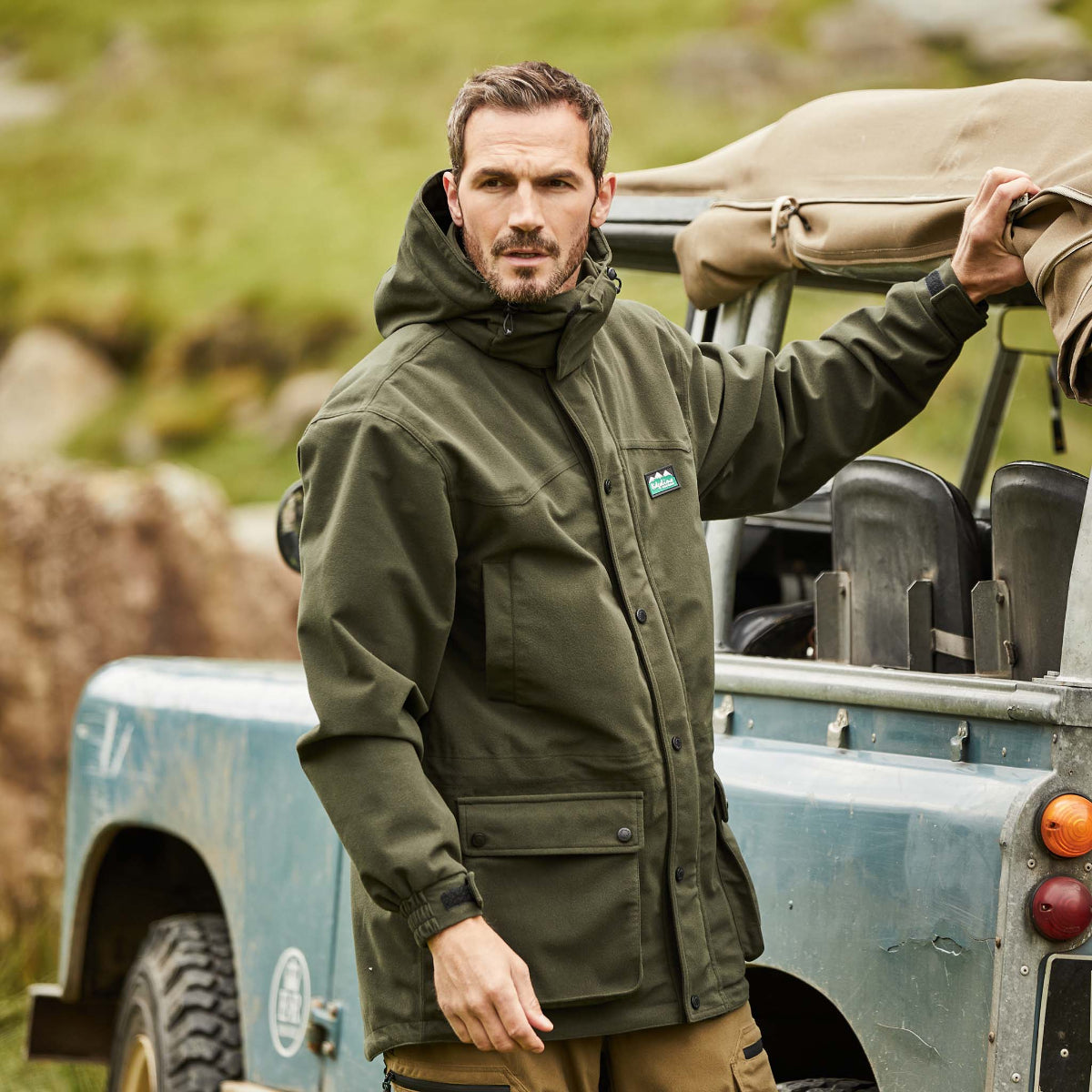 Man wearing a green-coloured Ridgeline Torrent III Jacket with a detachable hood, zip front, and multiple pockets. leaning on a jeep in an outdoor setting