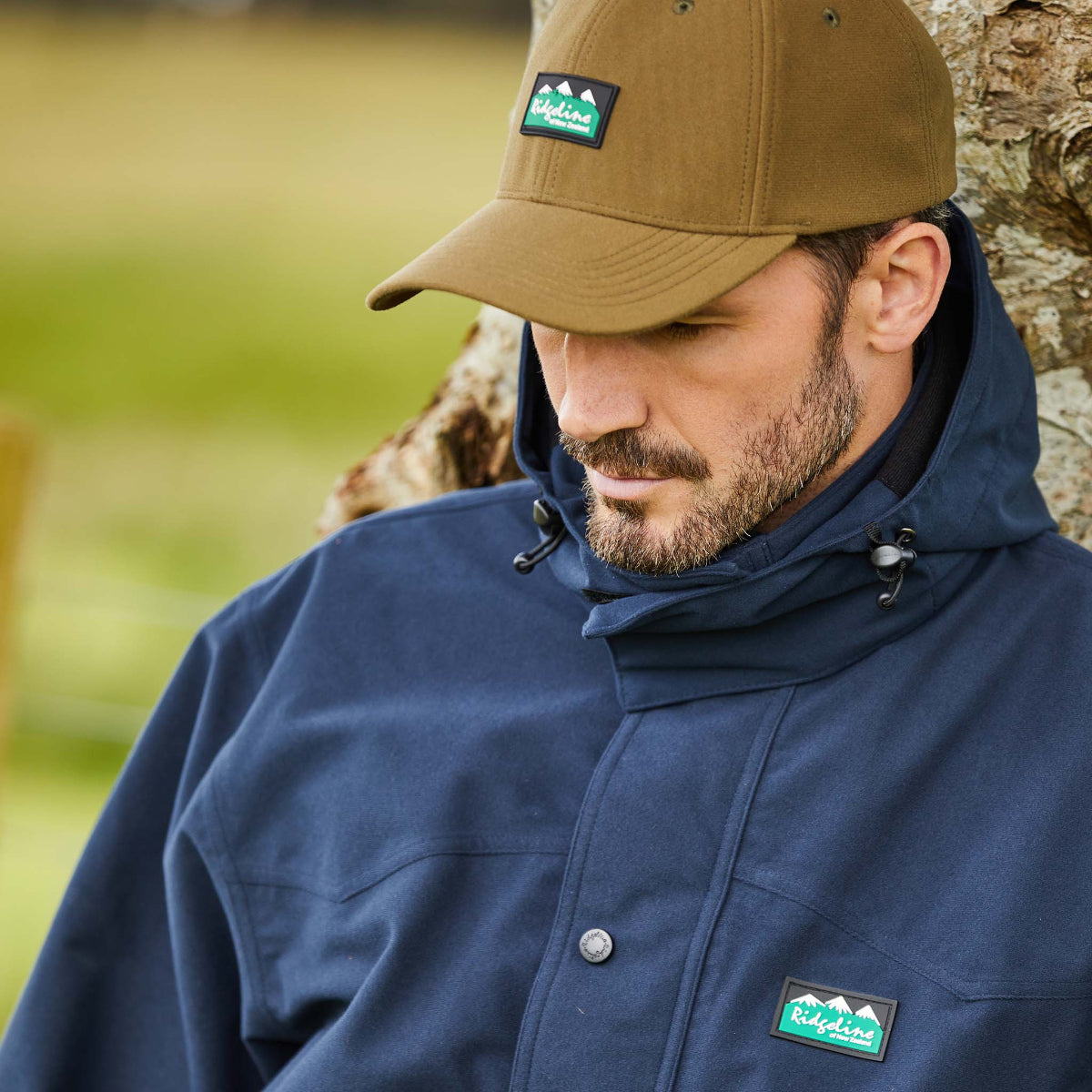 Man wearing a navy jacket and brown cap with a Ridgeline Clothing logo, sitting against a natural background.