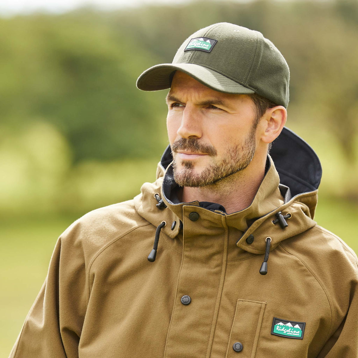 Man wearing a green Monsoon Classic Cap with an adjustable back and a curved peak, featuring a Ridgeline Clothing logo patch on the front. With a blurred outdoor background