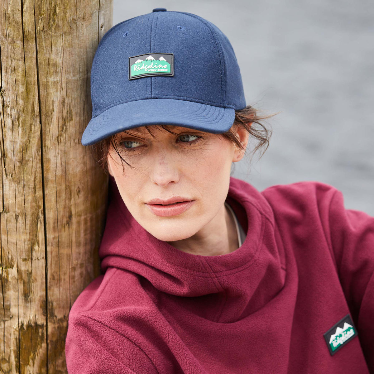 Lady wearing a Navy Ridgeline baseball cap with a curved peak and an adjustable back. Showing Ridgeline Clothing Logo on front. Leaning against a wooden plank 