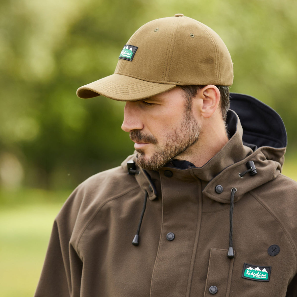 Man wearing a Brown Ridgeline baseball cap with a curved peak and an adjustable back. With a Ridgeline Clothing logo on front with a blurred outdoor background