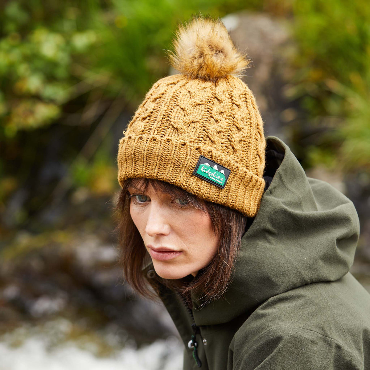 Woman wearing a brown Nordic Fleck bobble hat with a white pom-pom on top and a wide rib turn-up band. on a white background