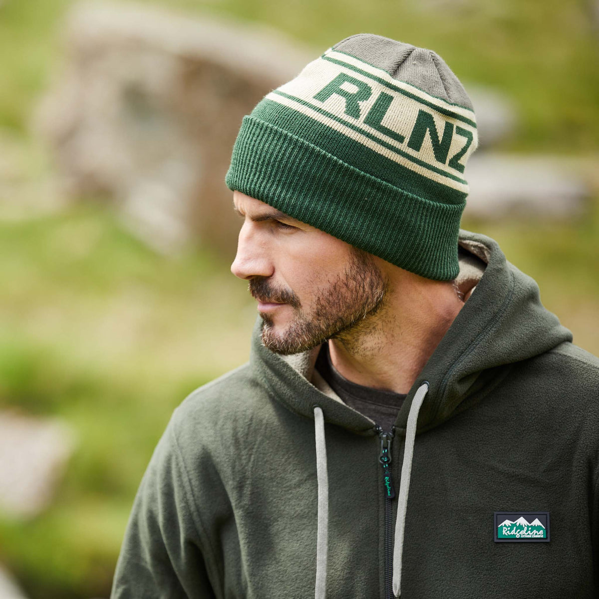 man wearing a knitted beanie with horizontal stripes in beige, green, and brown colours, featuring the text 'RLNZ' on the front. in an outdoor setting with blurred background