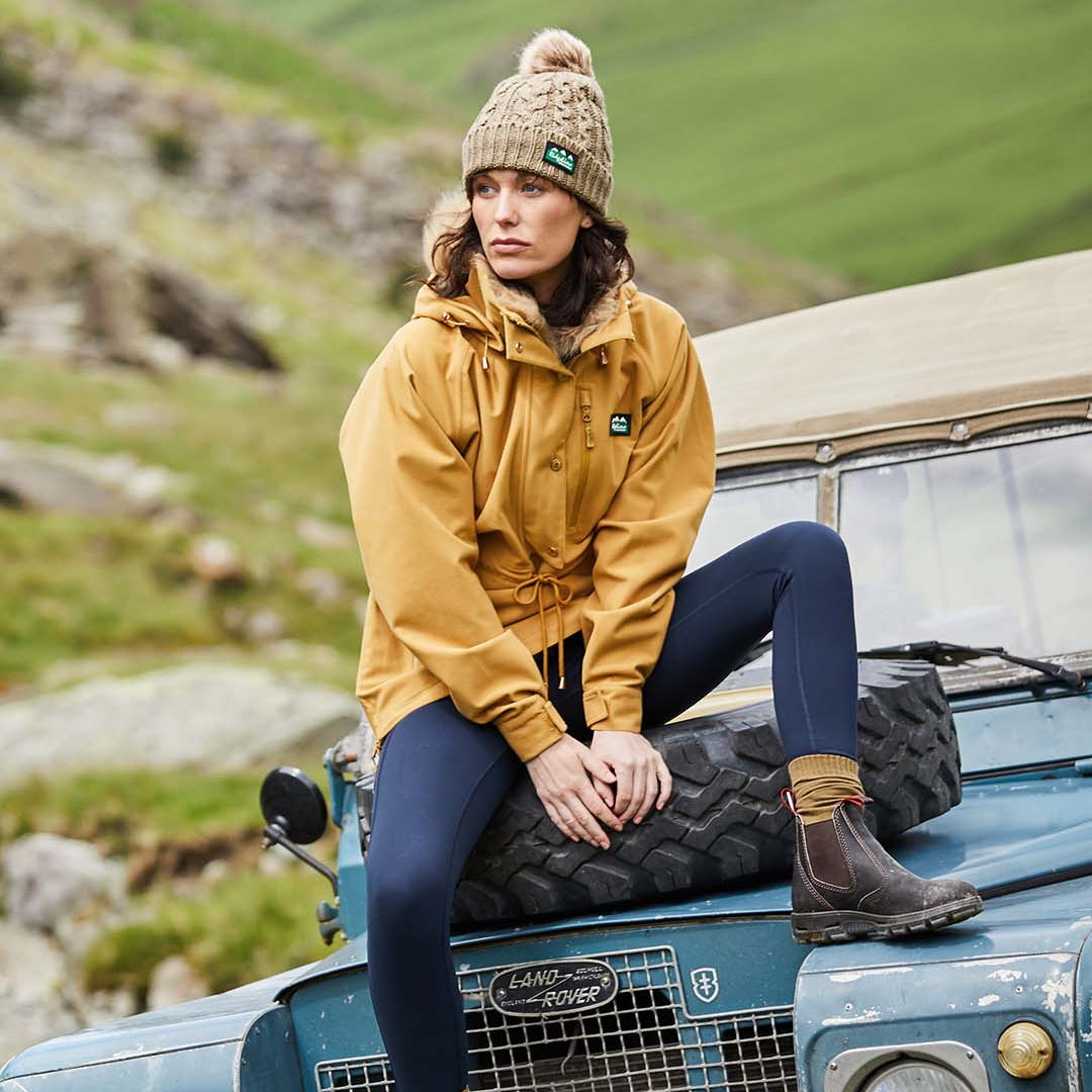 Woman in a yellow jacket and knit hat sitting on a Land Rover in a scenic outdoor setting.