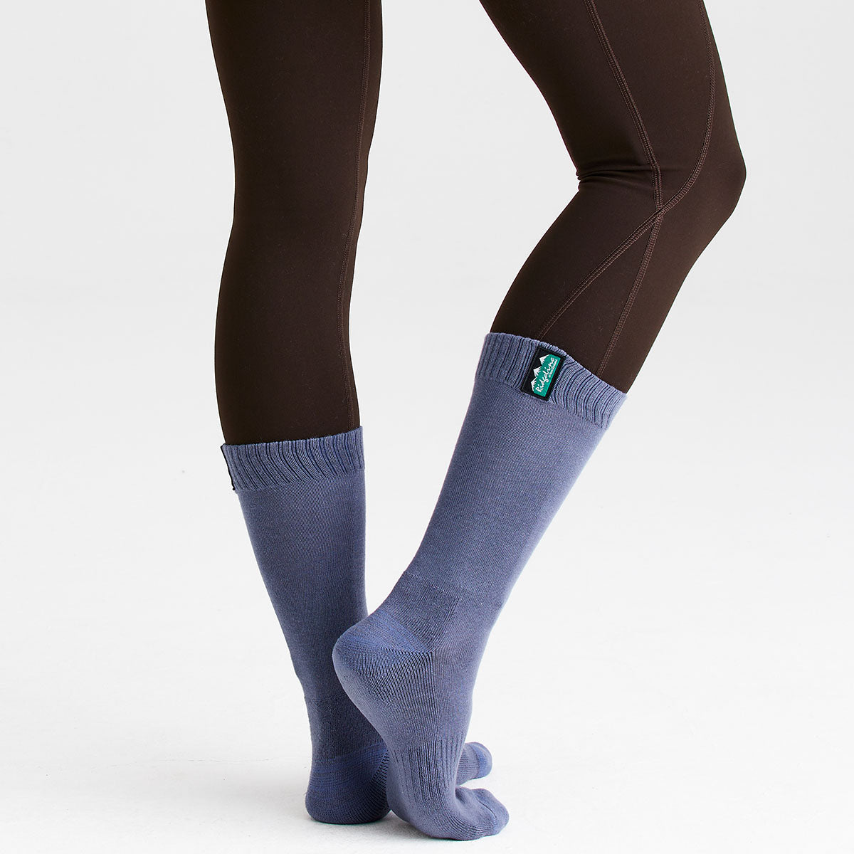 Person wearing light bluish grey full-length cotton work socks while standing on tiptoes with legs crossed against a light grey background. The knee-high socks feature a ribbed cuff and textured knit construction, with a visible Ridgeline logo tab near the top of one sock. The image showcases the fit, coverage, and comfort of the socks when worn.