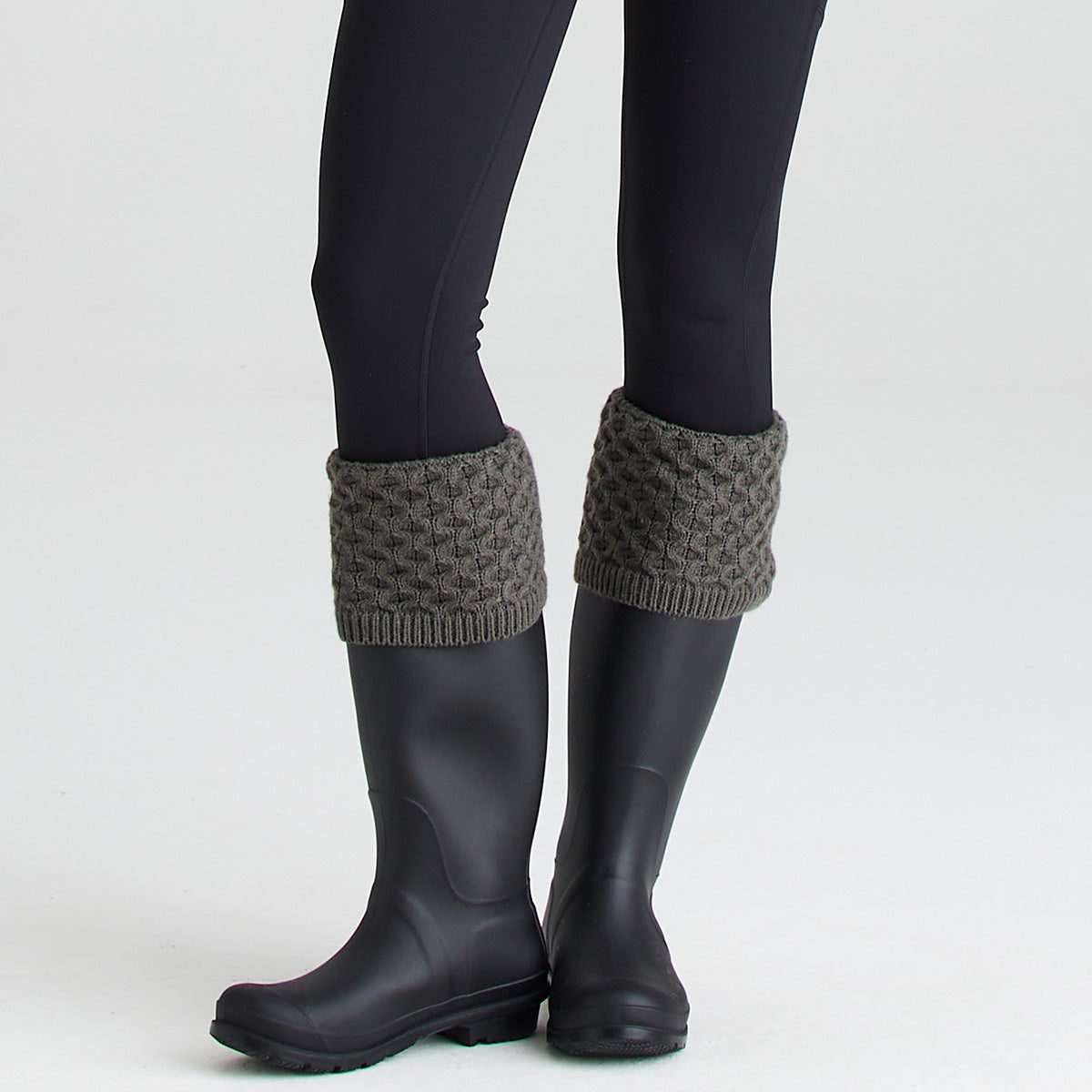 Close-up lifestyle image of a pair of dark rubber wellington boots worn with olive-green welly sock toppers folded over the boot openings. The welly socks feature a chunky cable-knit pattern with ribbed trim, creating a classic country-inspired look. The boots are paired with black leggings and photographed against a light grey studio background. The image highlights the knitted texture, comfortable fit, and added warmth and style the welly socks provide when worn with wellington boots.

