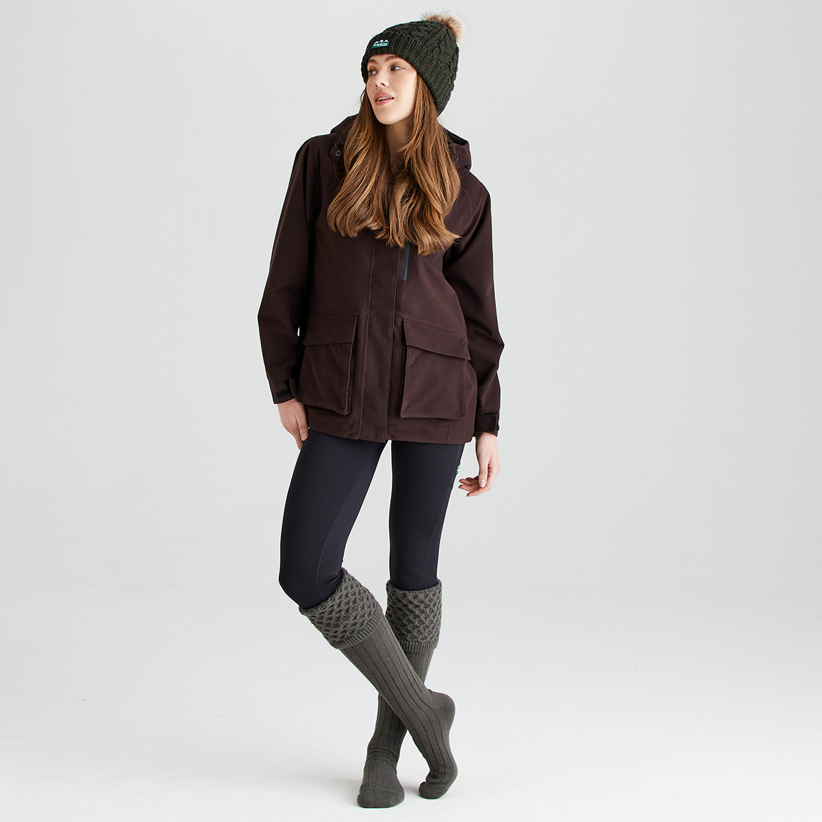 Lifestyle image showing a person wearing olive-green welly socks with a long ribbed leg and chunky cable-knit turnover cuffs. The socks are styled with black leggings, a dark outdoor jacket, and an olive-green cable-knit bobble hat featuring a faux-fur pom-pom and Ridgeline logo patch. Photographed against a light grey studio background, the image highlights the welly socks' classic country styling, knitted texture, and cosy fit as part of a coordinated outdoor outfit.
