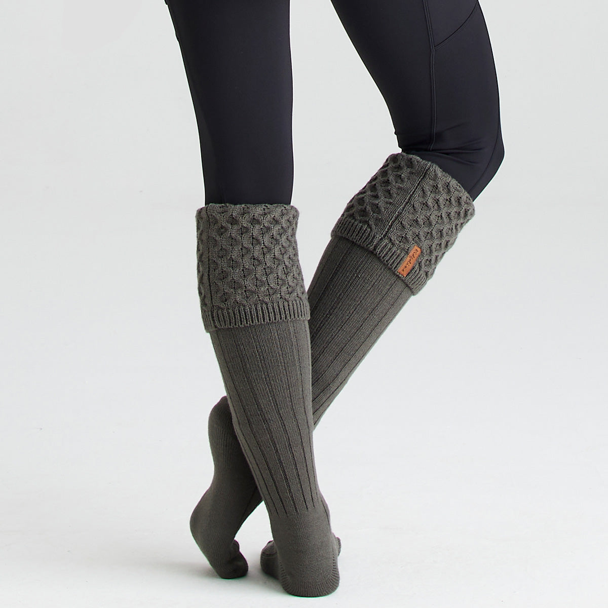 Lifestyle image of a person wearing olive-green welly socks with long ribbed legs and chunky cable-knit turnover cuffs. The socks are shown without boots and paired with black leggings, highlighting their knee-high length and textured knit construction. A small Ridgeline branded tag is visible on one cuff. Photographed against a light grey studio background, the image showcases the socks' cosy fit, classic country styling, and warm, comfortable design for outdoor wear.
